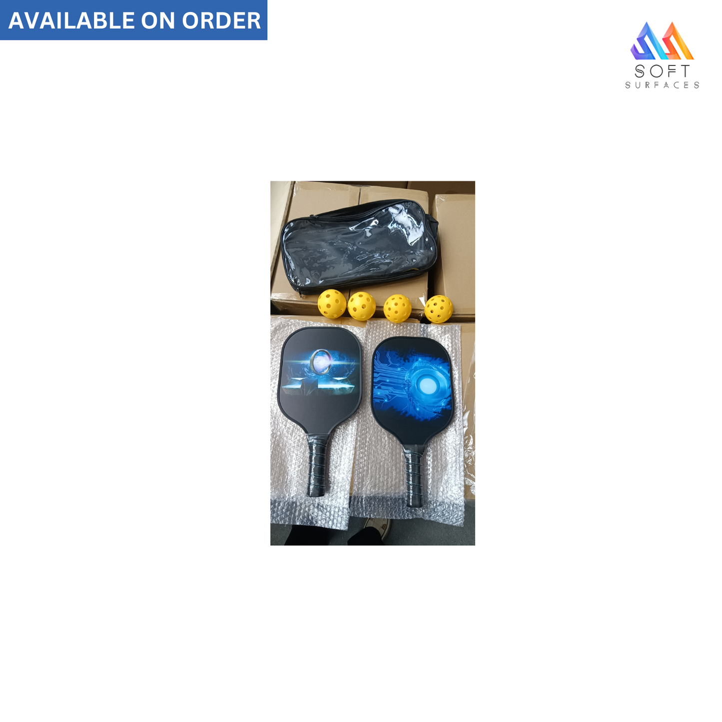 The image shows a set of pickleball rackets and balls. Two rackets with blue and black designs and one with white and black are visible alongside four yellow pickleballs.